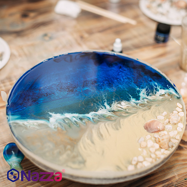 En la imagen se aprecia una pieza decorativa circular elaborada con resina epoxi de alto grosor, simulando un paisaje marino. La composición reproduce el efecto de una ola rompiendo sobre la arena: en la parte superior destacan intensos tonos azules y turquesa con matices blancos que imitan la espuma, mientras que en la inferior se observa una zona de arena con pequeñas piedras y conchas reales incrustadas. Todo está trabajado sobre una superficie de madera, transmitiendo un acabado brillante, nítido y de gran realismo, ideal para mostrar el potencial creativo de la resina epoxi en proyectos artísticos.